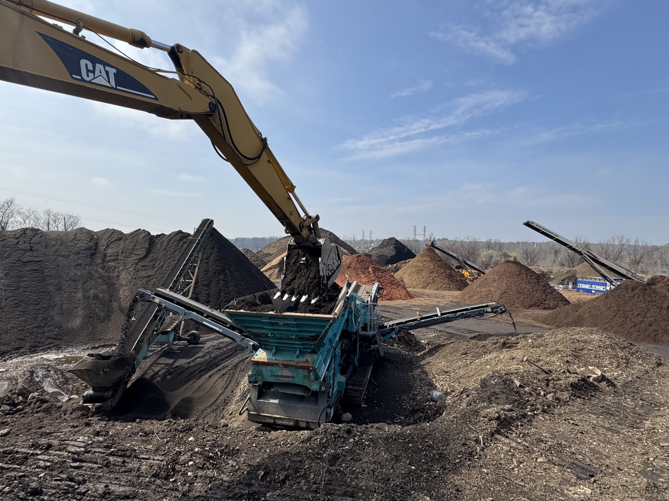 CAT excavator screening topsoil with bulk mulch operation visible in background at Grillo Services 20-acre facility in Milford CT